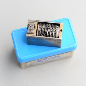 May include: A silver and brass number stamp with a black handle. The stamp has a series of rotating wheels with numbers 0-9 and a letter "N" on the side. The stamp is sitting on top of a blue plastic box with the word "Estonia" printed on it.
