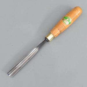 May include: A wood carving gouge with a light brown wooden handle and a metal blade. The handle has a brass ferrule and a green and white label. The blade is curved and has a polished finish.