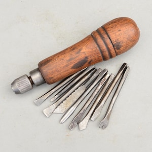 Könnte beinhalten: Ein Vintage-Handwerkzeug mit einem dunkelbraunen Holzgriff, einem strukturierten Metallfutter und neun austauschbaren Metallaufsätzen. Die Aufsätze haben verschiedene Formen, darunter Flachköpfe, spitze Enden und eine gezackte Klinge.