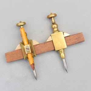 May include: Two antique brass and wood marking gauges. One gauge holds a pencil, the other has a metal point. Both are attached to a rectangular wooden beam. The brass components have a tarnished, aged appearance.