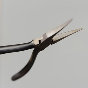 May include: Close-up of needle-nose pliers with a black handle and silver-colored metal jaws. The pliers are open, displaying the pointed tips. The tool is designed for precision work, likely for jewelry making or electronics.
