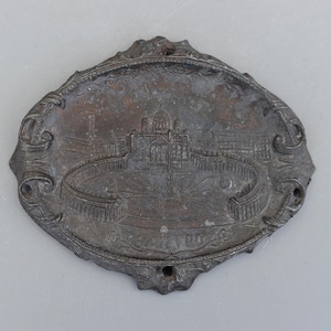 Puede incluir: Placa metálica ovalada de color plateado con un relieve detallado de un edificio con cúpula y columnata. La placa tiene volutas decorativas en los bordes y el texto "S. PIETRO".