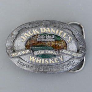 Könnte beinhalten: Ovale, silberfarbene Gürtelschnalle mit dem Jack Daniel's-Logo. Die Schnalle zeigt die Worte "Jack Daniel's" in Gold, mit "Old No. 7 Tennessee" und "Whiskey" ebenfalls in Gold. Sie lautet "Made with our own spring water".