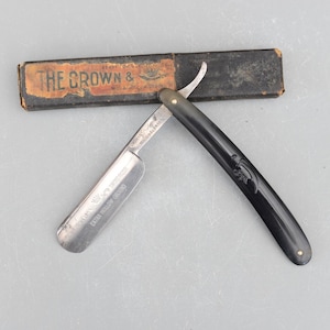 May include: An open straight razor with a black handle and a silver blade, next to a worn cardboard box. The box reads "THE CROWN &" in faded lettering. The razor blade is marked "EXTRA HOLLOW GROUND".