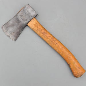 Puede incluir: Un hacha vintage con un mango de madera desgastado y una hoja de acero. El mango es de color marrón claro y la hoja muestra signos de desgaste. El hacha está diseñada para cortar madera y otras tareas.