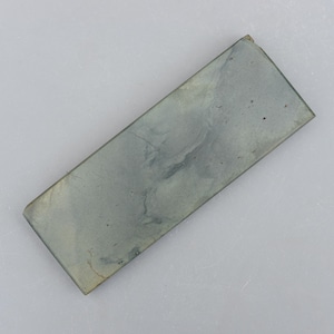 May include: A rectangular sharpening stone in a grey-green colour. The stone has a slightly textured surface with visible veining. The edges are slightly rounded, and the shape is a long rectangle. This tool is used for sharpening knives and other blades.