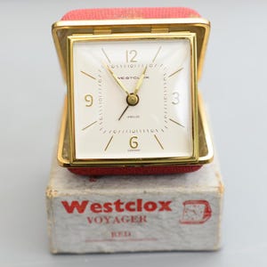 Puede incluir: Un reloj despertador de viaje Westclox Voyager vintage. El reloj cuadrado tiene un marco dorado y una esfera blanca con manecillas y números dorados. El reloj está en un estuche rojo y se encuentra en su caja original, que dice "Westclox Voyager RED".