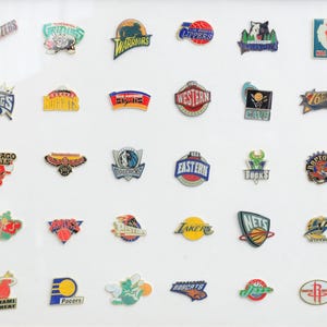 Puede incluir: Una colección de pines esmaltados con los logotipos de varios equipos de la NBA, incluidos los Lakers, Bulls y Warriors. Los pines son coloridos y detallados, mostrando los nombres y mascotas de los equipos. Los pines están dispuestos sobre una superficie blanca.