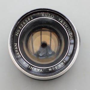 以下が含まれることがあります： 「AUTO YASHINON」と「JAPAN」の文字が黒とシルバーのレンズに印刷された、Yashinon 1:1.8 f=5.5cmカメラレンズ。レンズのシリアル番号はNo. 526586です。