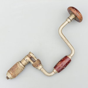 May include: A vintage hand drill with a metal frame and wooden handles. The drill has a curved design with a chuck at one end and a rotating handle at the other. The wooden handles are stained a dark reddish-brown colour.