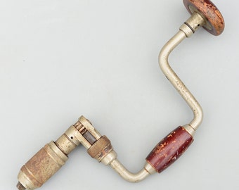 Op de afbeelding: Een vintage handboor met een metalen frame en houten handvatten. De boor heeft een gebogen ontwerp met een boorkop aan één uiteinde en een draaihendel aan de andere kant. De houten handvatten zijn donkerroodbruin gebeitst.