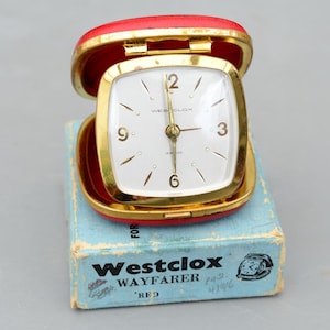 May include: A Westclox Wayfarer travel alarm clock with a red case and gold-tone trim, featuring a white square face with gold numerals and hands. The clock is displayed open, resting on its original light blue cardboard box with black text.