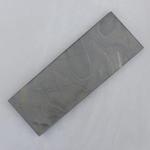 May include: A rectangular, gray sharpening stone with a marbled pattern. The stone has a smooth surface and is angled slightly, showcasing its rectangular shape. The stone's color varies from light to dark gray.