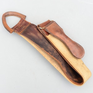 Peut inclure: Un cuir à rasoir vintage en cuir et toile. Il comporte une poignée en cuir marron avec le texte "SUPERIOR HIDE" et un corps en toile et cuir. Le cuir est conçu pour l'affûtage et l'entretien du tranchant des lames.