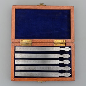 May include: A wooden box with a blue velvet lining contains five metal rulers. Each ruler is marked with inch measurements and has a rounded end with text. The box is made of wood and has brass hinges.