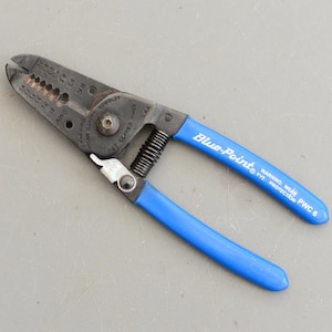 Puede incluir: Pelacables Blue-Point con mangos negros y empuñaduras azules. La herramienta tiene marcas para diferentes calibres de cables y una advertencia para usar protección para los ojos.