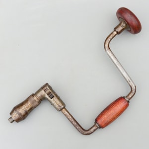 May include: A vintage hand drill with a wooden handle and a metal crank. The drill is made of metal and has a rusty patina. The handle is made of wood and has a dark brown finish.