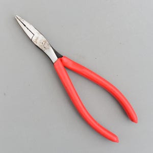 Könnte beinhalten: Eine Spitzzange mit einem silberfarbenen Metallkopf und leuchtend roten Griffen. Die Zange ist geöffnet und zeigt die spitzen Spitzen. Das Werkzeug dient zum Greifen, Biegen und Schneiden kleiner Objekte.