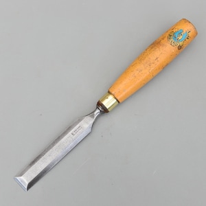 May include: A wood chisel with a light orange wooden handle and a silver blade. The handle has a brass ferrule and a blue and gold logo. The blade is rectangular and has text on it. The tool is designed for woodworking.