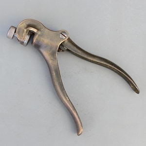 May include: A vintage-style metal hand tool with a dark, weathered bronze finish. The tool features two curved handles, a central pivot point secured by a flat-head screw, and an adjustable circular mechanism at the top with a knurled metal knob.