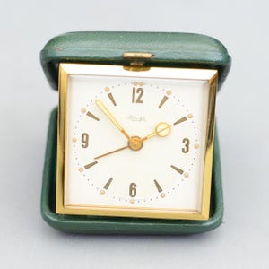 以下が含まれることがあります： 金色の正方形のフレームと白い文字盤を備えた、ヴィンテージの旅行用目覚まし時計。 時計には黒い数字と金の針があり、緑色の合皮ケースに収められています。 時計の文字盤には「Kiengle」と書かれています。