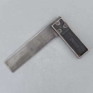 May include: A vintage metal try square with a dark finish. The ruler arm is marked with inch measurements from 1 to 6. The handle is black with the word "RABONE" embossed. A classic tool for woodworking and metalworking.