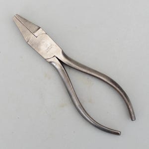 Può includere: Un paio di pinze in metallo argentato con manici lunghi e curvi. Le pinze hanno un naso piatto e appuntito e una cerniera al centro. La parola "PEREZ" è stampata sul lato delle pinze.