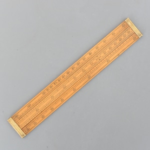 以下が含まれることがあります： 真鍮のエンドピースが付いた、ヴィンテージの木製計算尺。計算尺には、計算用の数字や線など、複数の目盛りとマーキングがあります。木材は明るい茶色で、真鍮は金色です。計算尺の長さは約30cmです。