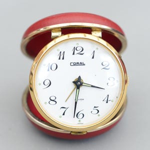 以下が含まれることがあります： 白い文字盤と黒い数字の金色の目覚まし時計。時計は赤い丸いケースに入っています。時計の文字盤には「CORAL」、「ARM」、「JAPAN」と書いてあります。
