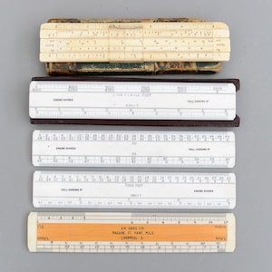 Puede incluir: Una colección de reglas de cálculo antiguas, incluyendo una beige con estuche de cuero y otra de madera con el texto "A.H. Davis Ltd, Liverpool 3". Estas herramientas se usaban para cálculos matemáticos.