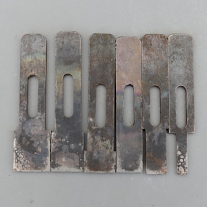 Może przedstawiać: Sześć metalowych elementów w stylu vintage o prostokątnym kształcie i podłużnym owalnym wycięciu. Metal ma zużyty, zmatowiony wygląd z przebarwieniami. Elementy ułożone są obok siebie na szarej powierzchni.