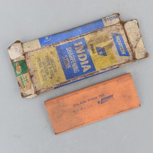 May include: An open, weathered box with blue and yellow text reading "INDIA SHARPENING STONE" next to an orange sharpening stone. The box also has text in multiple languages. The orange stone has the text "Fine India Oilstone FS44" and "MADE IN USA".