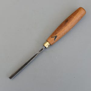 Puede incluir: Un formón de carpintería con mango de madera y hoja de metal. El mango es de color marrón claro con una flecha azul oscuro. La hoja metálica es plateada y tiene un borde plano. Herramienta para tallar y dar forma a la madera.