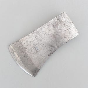 May include: A weathered steel axe head with a curved blade and a slightly textured surface. The axe head has a rectangular shape with rounded corners and a small, faded marking near the centre. The metal has a dull, grey appearance.