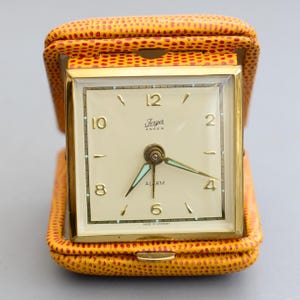 Könnte beinhalten: Ein Vintage-Reisewecker mit cremefarbenem Zifferblatt, goldfarbenem Rahmen und türkisfarbenen Zeigern. Er befindet sich in einem gelben Lederetui mit rotem Punktmuster. Das Zifferblatt trägt die Marke "Ferger ANKER" und das Wort "ALARM".