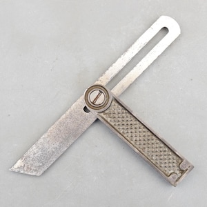 May include: A vintage metal sliding bevel gauge, a woodworking tool, with a textured handle and a pivoting blade. The blade is open, showing the angle. The tool is made of metal and has a worn, used appearance.