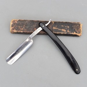 Puede incluir: Una navaja de afeitar recta vintage con una hoja plateada y un mango negro. La hoja tiene texto y el mango es curvo. Se encuentra junto a un estuche de cuero marrón desgastado. Una herramienta de aseo personal.