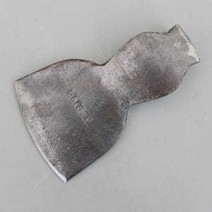 May include: A worn steel axe head with a curved blade and a flared top. The metal has a dark, aged appearance, with the word "ZARAMA" stamped on the surface. The axe head is set against a grey background.