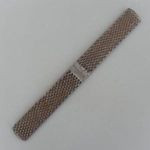 May include: A metal rasp with a rectangular shape and a textured surface. The rasp is a tool used for shaping wood or other materials. The surface is covered in small, closely spaced teeth.