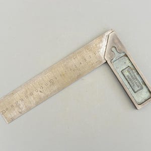 May include: A silver metal try square with a ruler marked in inches from 2 to 20. The try square has a label that reads "Sugavu" in all capital letters.