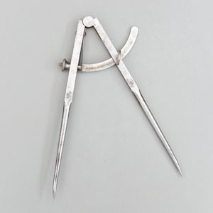 以下が含まれることがあります： 2 つの尖った脚と調整ネジが付いた、ヴィンテージの金属製コンパス。コンパスは銀色で、鋼製と思われます。距離を測定したり、円を描いたりするためのツールです。