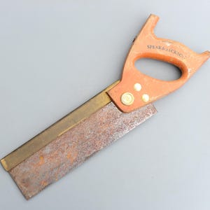以下が含まれることがあります： 真鍮の刃とオレンジ色のプラスチック製のハンドルが付いたビンテージの手のこぎり。ハンドルには「Spear & Jackson」の文字が印刷されています。