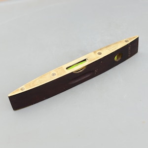 May include: A vintage spirit level with a dark wood body and brass edges. The level has a small, curved glass vial filled with a green liquid and is approximately 12 inches long. The brass is engraved with text.