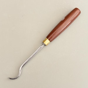 Puede incluir: Una herramienta para trabajar la madera con una hoja metálica curva y un mango de madera marrón oscuro. El mango está unido a la hoja con una férula de latón. La herramienta está diseñada para tallar o dar forma a la madera.