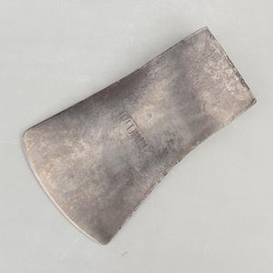 May include: A weathered, rectangular metal axe head with a curved edge. The axe head has a silver finish and a slightly textured surface. The word "PLUMB" is stamped into the metal. The axe head is designed for woodworking and carpentry.