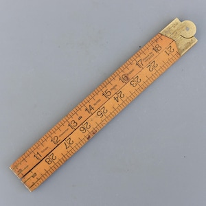 Puede incluir: Una regla plegable de madera vintage con accesorios de latón. La regla está marcada con medidas en pulgadas y números. El texto "Chesterman" y "Patent No 1173229" son visibles. La regla es de color marrón claro.