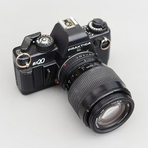 Peut inclure: Un appareil photo argentique noir Praktica BX20 avec un objectif Praktica 1.2/24-70 MC Macro. L'appareil photo a une bague argentée autour de l'objectif et une bague argentée autour du corps.