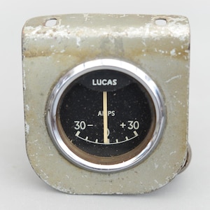Peut inclure: Un indicateur automobile argenté avec un cadran noir et une lunette chromée. L'indicateur affiche "Lucas" et "AMPS" et a une aiguille pointant vers zéro. L'indicateur est monté sur un support en métal argenté.