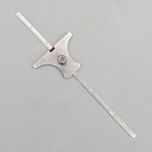 Op de afbeelding: Een vintage metalen dieptemeter met een schuifschaal en een centrale afstelknop. De meter heeft een plat, vleugelvormig lichaam en twee uitstekende armen met meetmarkeringen. De tool is ontworpen voor precieze dieptemetingen.