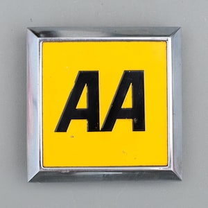 May include: A square metal badge featuring a bright yellow centre panel with the bold black letters AA printed in the middle. The emblem is framed by a polished silver-coloured metallic border with beveled edges, creating a clean and distinct look.